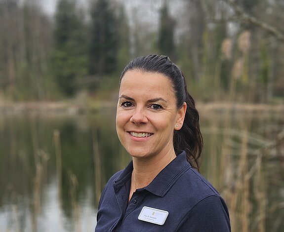 Die stellvertretende Therapieleitung der KIH in dunklem Poloshirt mit Namensschild steht lächelnd vor einem See in natürlicher Umgebung.