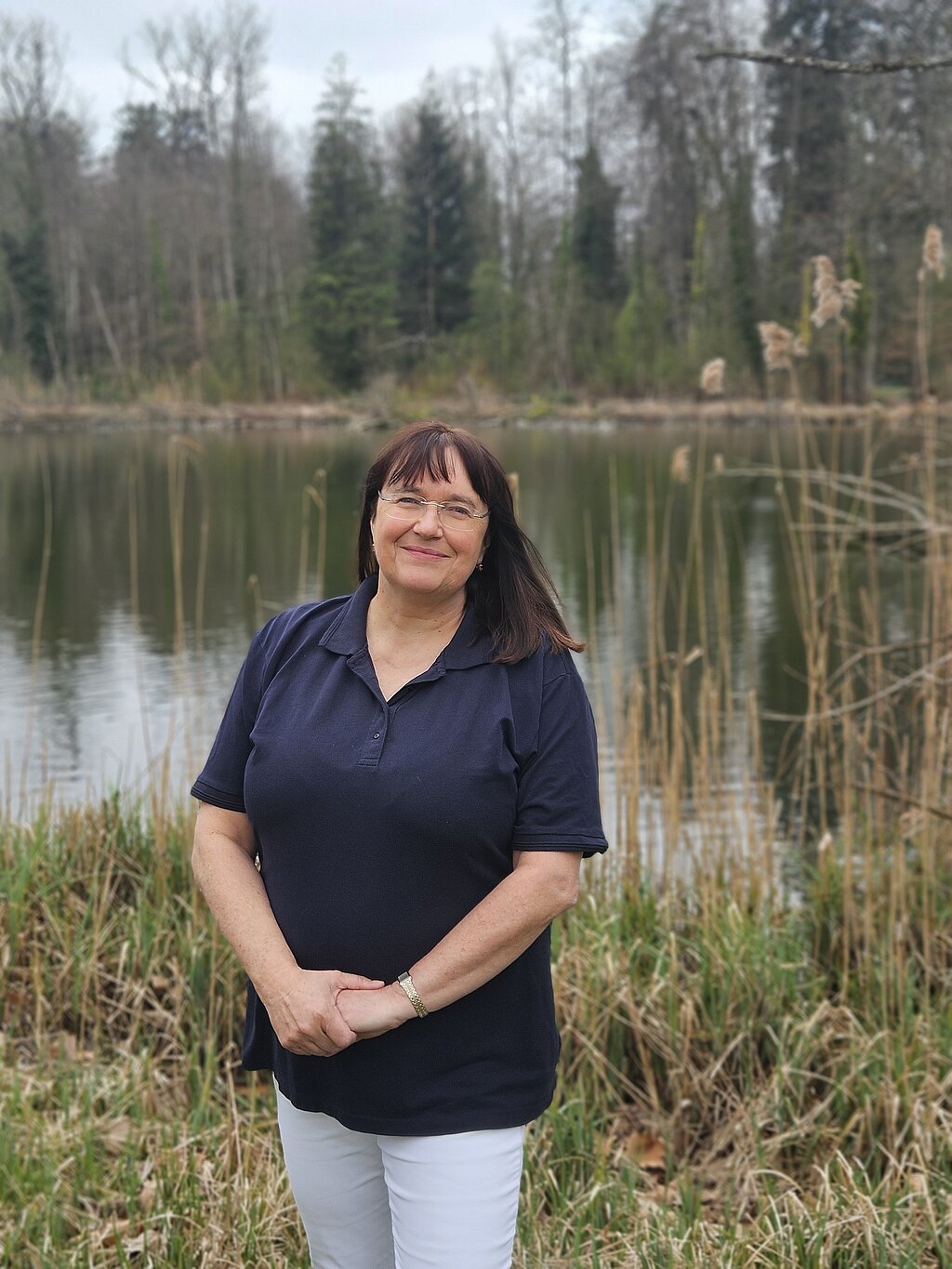 Die Therapieleitung der KIH steht in dunklem Shirt vor einem See und lächelt Richtung Kamera , im Hintergrund Bäume und Natur.