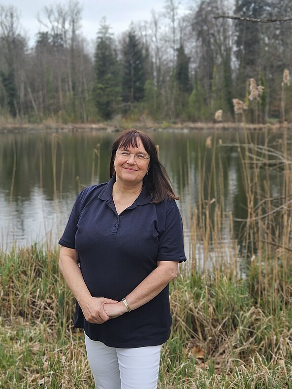 Die Therapieleitung der KIH steht in dunklem Shirt vor einem See und lächelt Richtung Kamera , im Hintergrund Bäume und Natur.