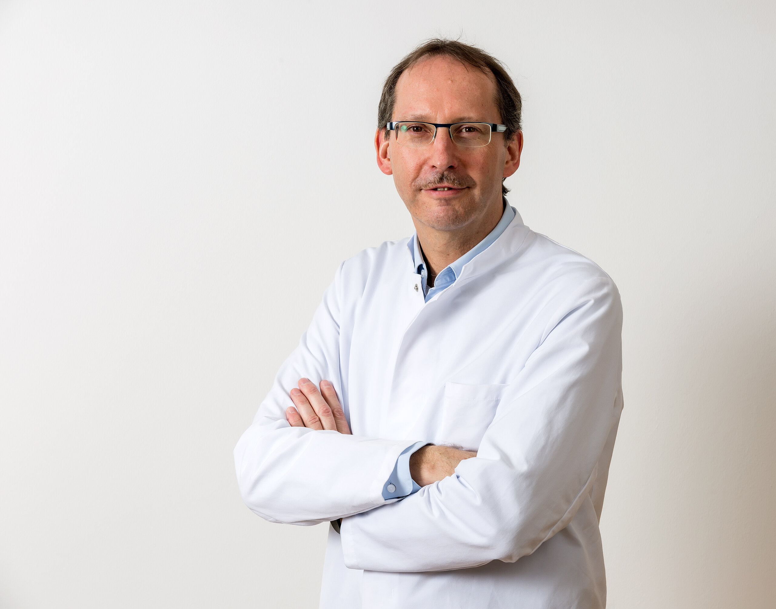 Dr. Klaus Müller, Chefarzt der Klinik im Hofgarten, steht im weißen Arztkittel mit verschränkten Armen vor hellem Hintergrund und blickt freundlich in die Kamera.