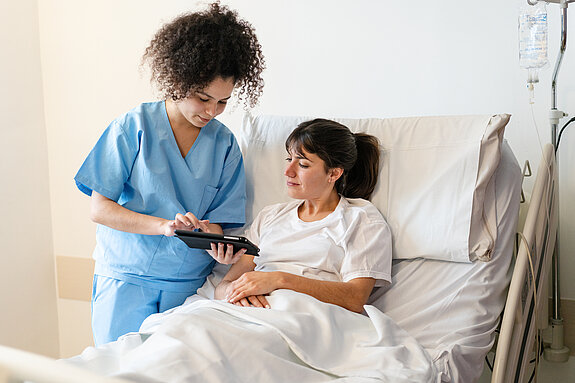 Eine Pflegekraft in blauer Kleidung zeigt einer Patientin im Krankenhausbett etwas auf einem Tablet.