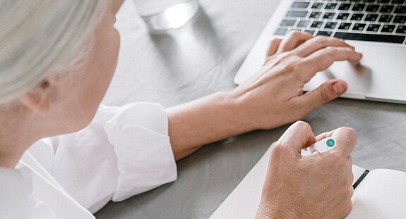 Ältere Frau in weißem Kittel sitzt an einem Schreibtisch, schreibt auf Papier und arbeitet an einem Laptop.