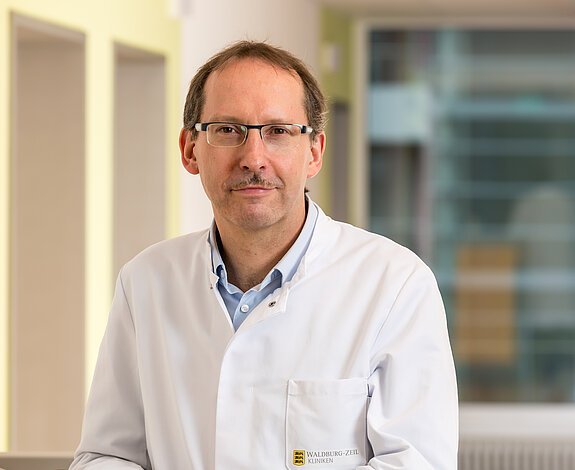 Dr. Klaus Müller, Chefarzt der Klinik im Hofgarten, steht an einem Stationswagen und lächelt freundlich