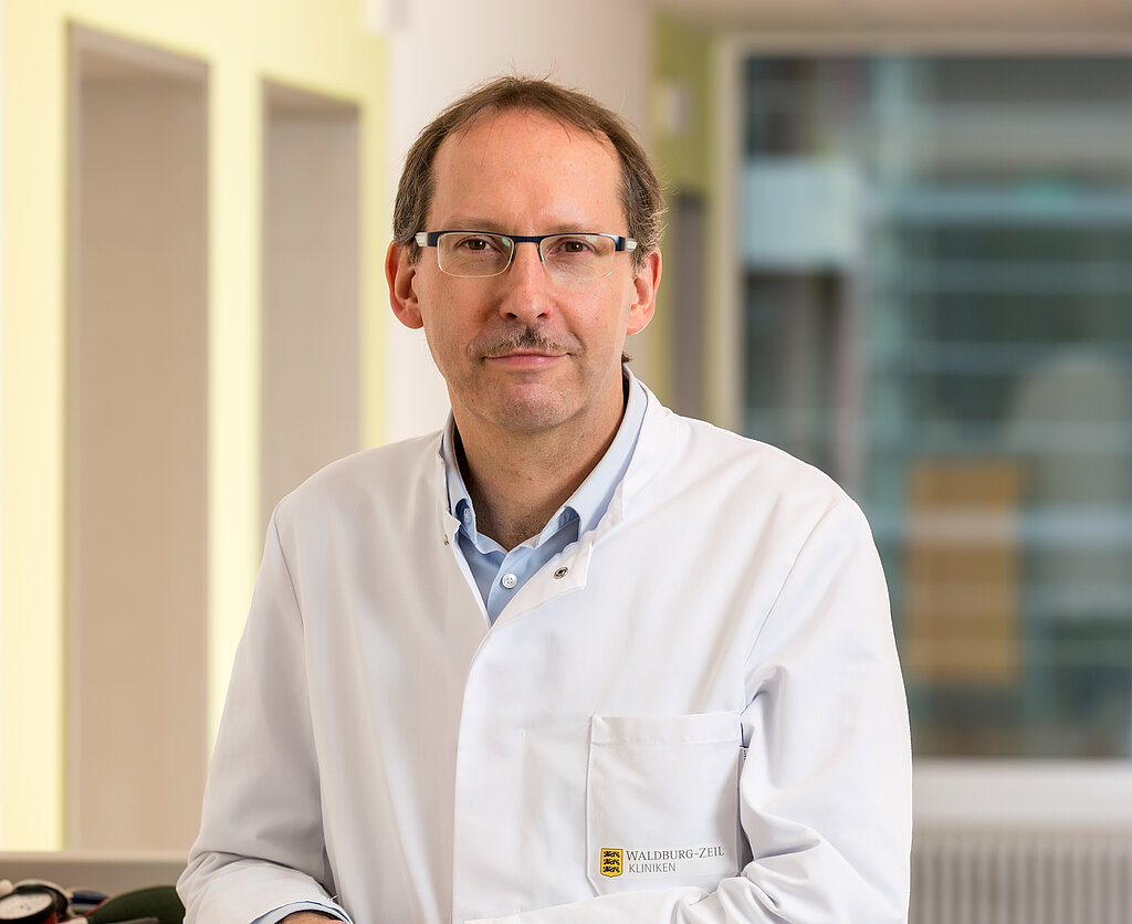 Dr. Klaus Müller, Chefarzt der Klinik im Hofgarten, steht an einem Stationswagen und lächelt freundlich
