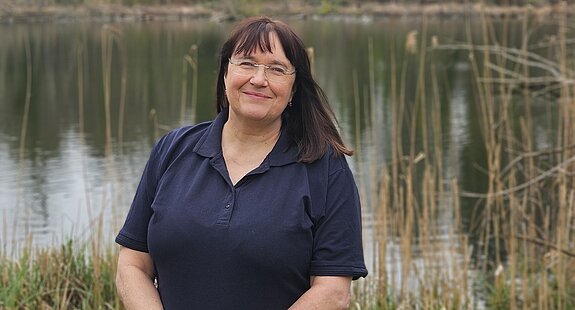 Die Therapieleitung der KIH steht in dunklem Shirt vor einem See und lächelt Richtung Kamera , im Hintergrund Bäume und Natur.
