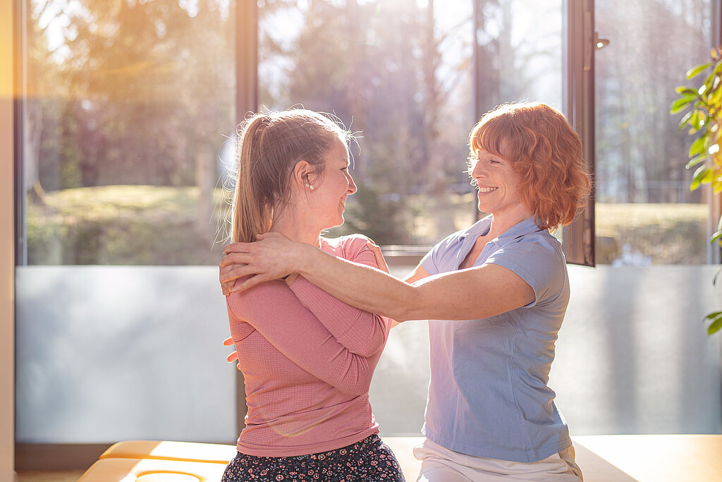 Zwei Frauen in einem hellen Raum führen eine physiotherapeutische Übung am Oberkörper durch und lächeln sich an