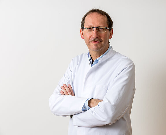 Dr. Klaus Müller, Chefarzt der Klinik im Hofgarten, steht im weißen Arztkittel mit verschränkten Armen vor hellem Hintergrund und blickt freundlich in die Kamera.