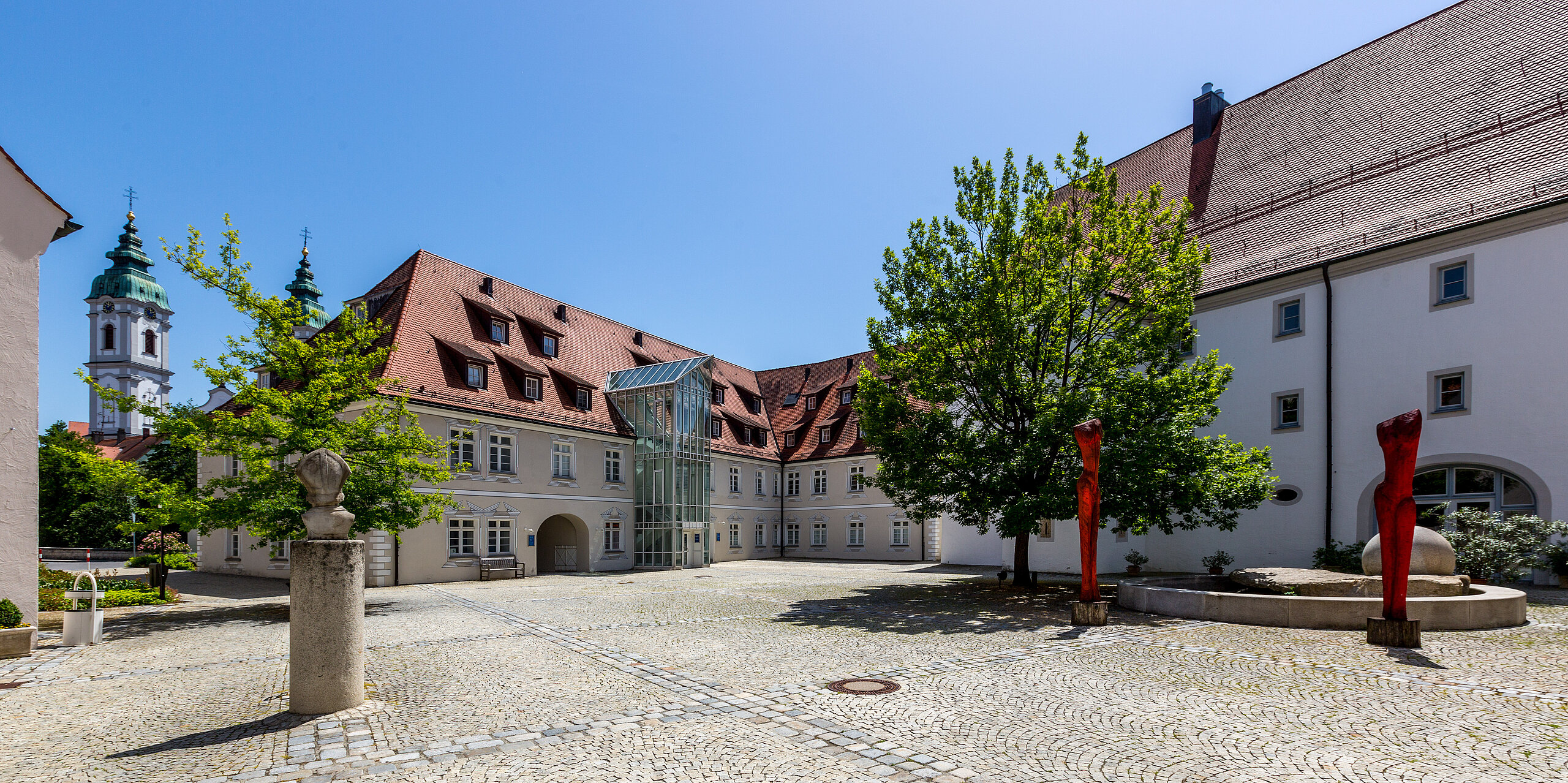 Gepflasterter Innenhof der Klinik im Hofgarten in Bad Waldsee, umgeben von historischen Gebäuden mit roten Dächern, Bäumen und blauem Himmel im Hintergrund