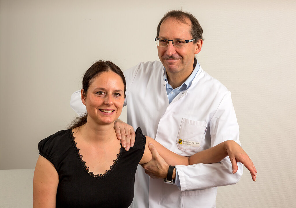 Dr. Klaus Müller, Chefarzt der Klinik im Hofgarten, steht im weißen Arztkittel mit verschränkten Armen vor hellem Hintergrund und blickt freundlich in die Kamera.